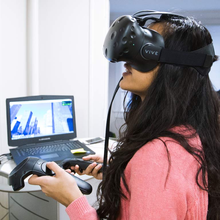 A student uses a virtual reality headset and controllers while a laptop shows what she is seeing.