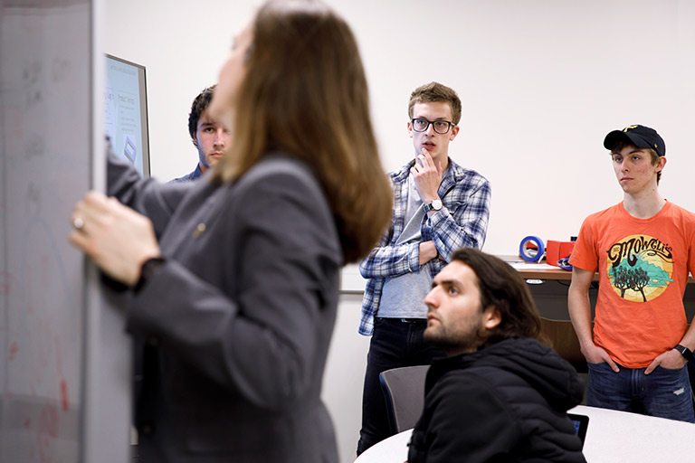 A professor writes notes on a whiteboard as a small group of students watch.